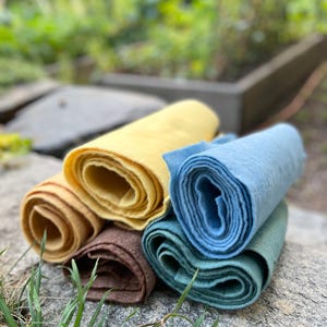 Op de afbeelding: Een verzameling opgerolde stoffen in verschillende kleuren, waaronder geel, bruin, blauw en groen. De stoffen liggen op een grijze stenen ondergrond, met een vage groene achtergrond.