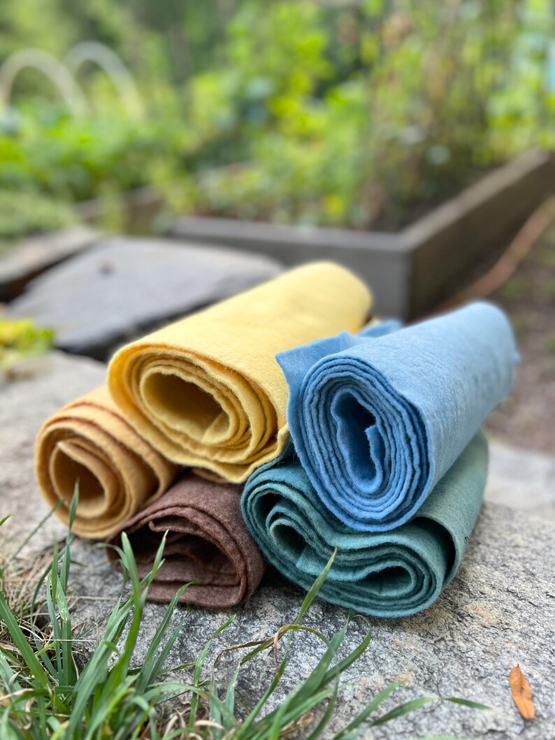 Op de afbeelding: Een verzameling opgerolde stoffen in verschillende kleuren, waaronder geel, bruin, blauw en groen. De stoffen liggen op een grijze stenen ondergrond, met een vage groene achtergrond.