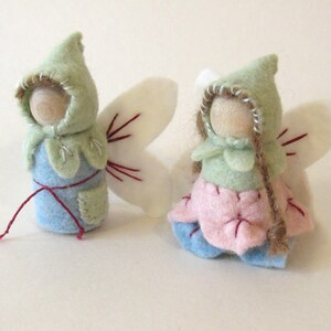 May include: Two small peg dolls with wooden heads and felt clothing. One doll wears a light blue felt body with a green hood and white wings. The other doll wears a pink petal-shaped skirt, a green hood, and white wings with brown yarn hair.