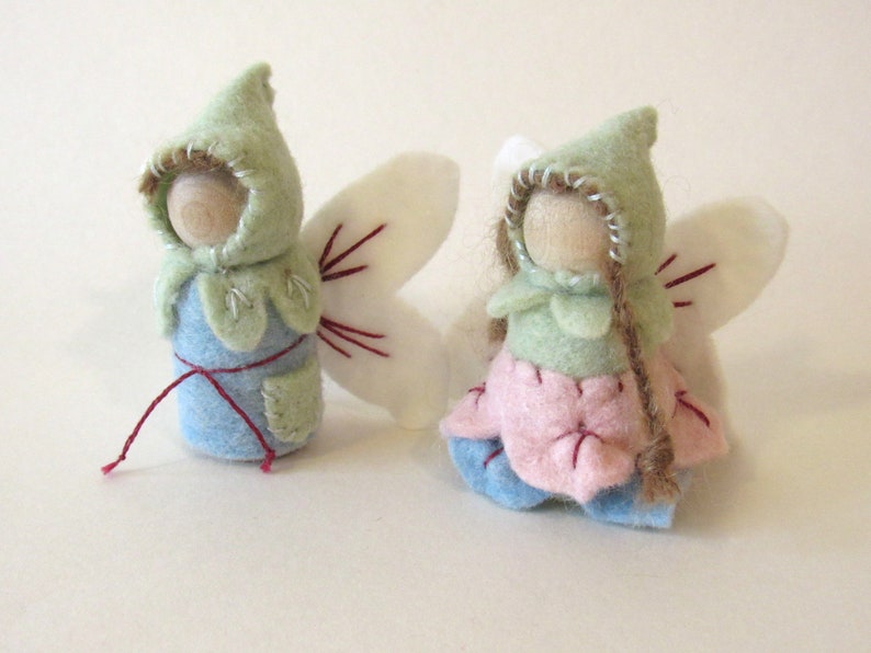May include: Two small peg dolls with wooden heads and felt clothing. One doll wears a light blue felt body with a green hood and white wings. The other doll wears a pink petal-shaped skirt, a green hood, and white wings with brown yarn hair.
