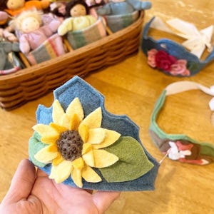 Op de afbeelding: Een blauwe vilten kroon versierd met een gele zonnebloem en groene bladeren. Andere vilten kronen en poppen in een mand zijn op de achtergrond. De kroon wordt in een hand gehouden.