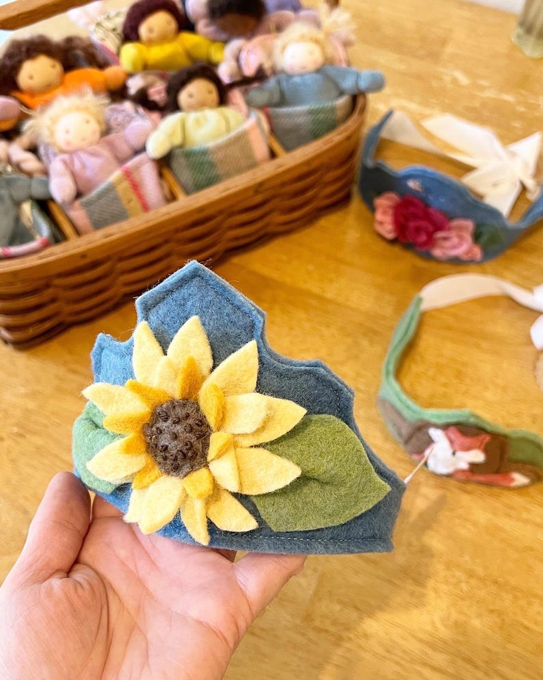 Op de afbeelding: Een blauwe vilten kroon versierd met een gele zonnebloem en groene bladeren. Andere vilten kronen en poppen in een mand zijn op de achtergrond. De kroon wordt in een hand gehouden.