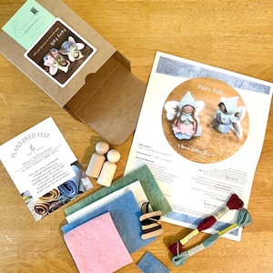 May include: A craft kit for making felt fairy dolls, including a cardboard box, two wooden peg dolls, sheets of pink, blue, and green felt, two skeins of embroidery floss, a small wooden spool with thread, a needle, and an instructional guide.