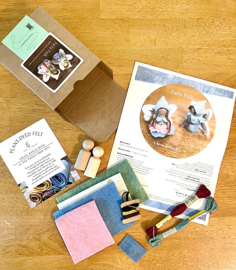 May include: A craft kit for making felt fairy dolls, including a cardboard box, two wooden peg dolls, sheets of pink, blue, and green felt, two skeins of embroidery floss, a small wooden spool with thread, a needle, and an instructional guide.