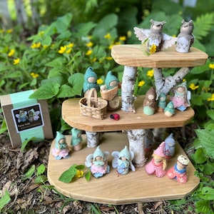 May include: A three-tiered wooden display stand made of birch branches and smooth wood platforms, holding numerous small felted figures including owls, fairies, and gnomes. A cardboard box labeled Fairy Folk sits to the left of the display.