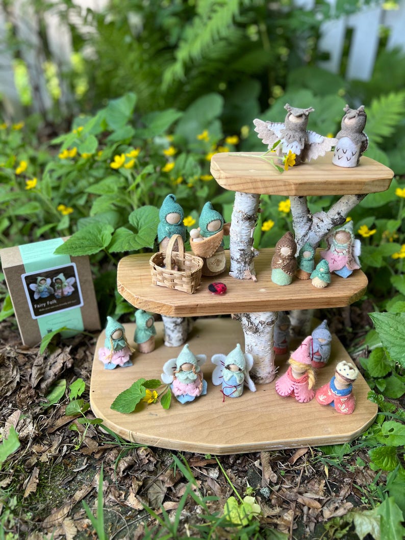 May include: A three-tiered wooden display stand made of birch branches and smooth wood platforms, holding numerous small felted figures including owls, fairies, and gnomes. A cardboard box labeled Fairy Folk sits to the left of the display.