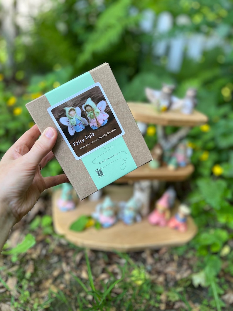 May include: A rectangular brown cardboard box with a light green paper band featuring a white label. The label displays two small felt fairy figures, the text Fairy Folk, Make your own eco-friendly felt toys, and A hand-sewing project.
