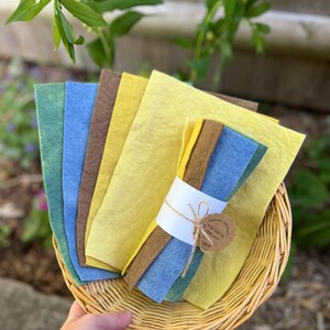 Op de afbeelding: Een hand houdt een gevlochten mand met gekleurde vellen vilt. Het vilt is in tinten groen, blauw, bruin en geel. Een bundel vilt is verpakt met een witte band en vastgebonden met touw, met een klein label met de tekst "WEIR THINGS CRAFT ARTS".