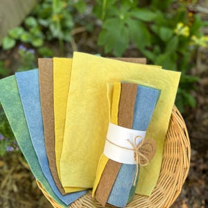 Op de afbeelding: Een verzameling kleurrijke vellen vilt in tinten geel, blauw, groen en bruin, gerangschikt in een rieten mand. Een bundel vilt is vastgebonden met touw en een label. Het vilt wordt buiten tentoongesteld.