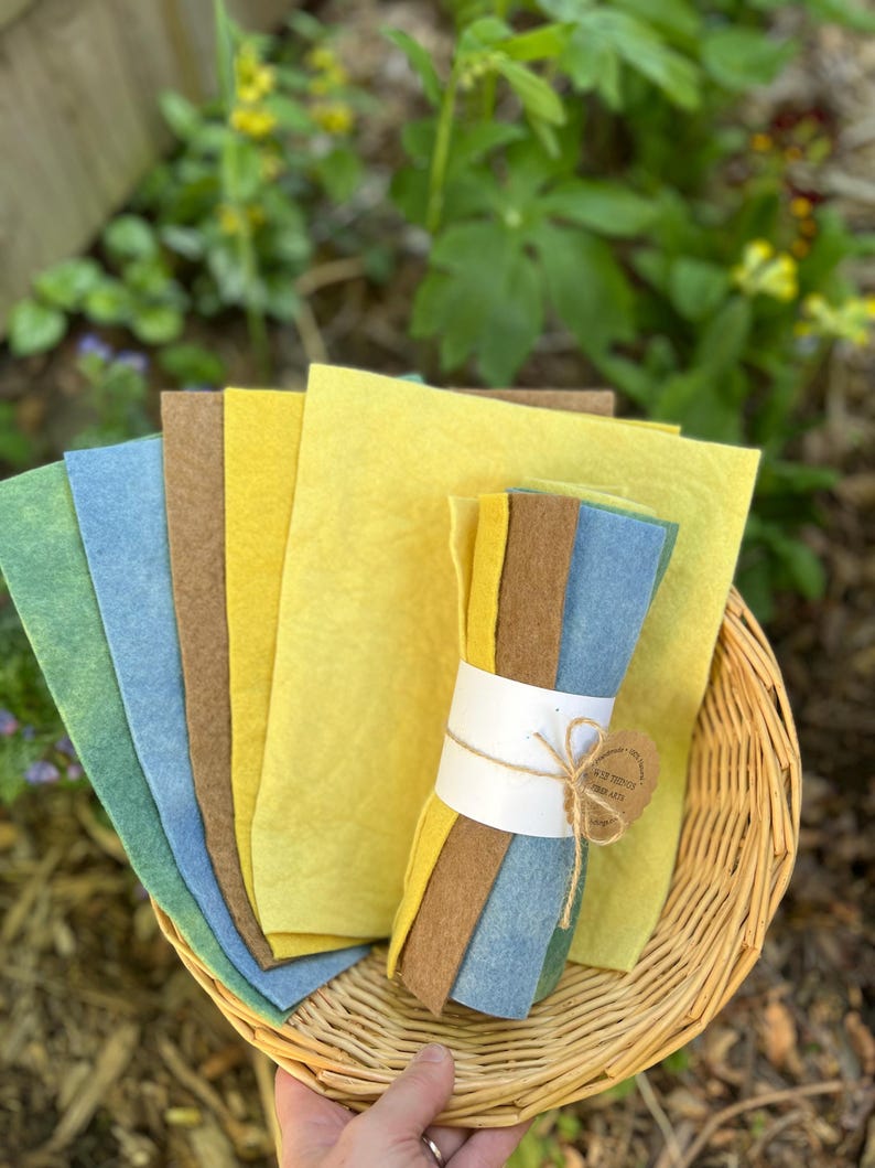 Op de afbeelding: Een verzameling kleurrijke vellen vilt in tinten geel, blauw, groen en bruin, gerangschikt in een rieten mand. Een bundel vilt is vastgebonden met touw en een label. Het vilt wordt buiten tentoongesteld.