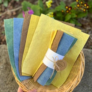 Op de afbeelding: Een verzameling kleurrijke vellen vilt in groen, blauw, bruin en geel, gerangschikt in een rieten mand. Een bundel vilt is verpakt met een witte band en vastgebonden met touw, met een klein label. De achtergrond toont groen en bloemen.