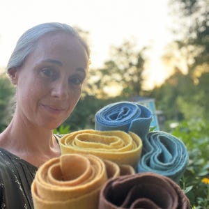 Op de afbeelding: Een close-up van opgerold vilt in verschillende kleuren: lichtbruin, geel, blauw en turquoise. Het vilt is strak opgerold, waardoor de textuur en kleurvariaties zichtbaar zijn. De achtergrond is een vage buitensc&egrave;ne.