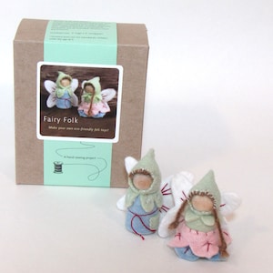 May include: A craft kit box labeled Fairy Folk with a light green band and two small handmade felt fairy dolls. The dolls feature wooden peg heads, pointed green hoods, white wings, and skirts in blue and pink with brown yarn hair details.