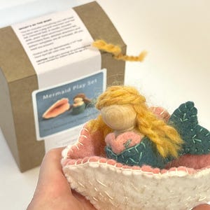 May include: A felt mermaid doll with yellow yarn hair and a teal tail, resting inside a white and pink felt shell. The mermaid has a wooden bead head. In the background, a brown cardboard box features a label with the text Mermaid Play Set.