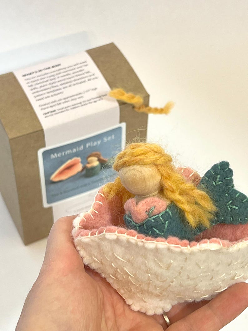 May include: A felt mermaid doll with yellow yarn hair and a teal tail, resting inside a white and pink felt shell. The mermaid has a wooden bead head. In the background, a brown cardboard box features a label with the text Mermaid Play Set.