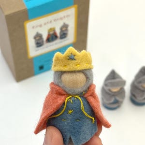 May include: A felt king figurine featuring a yellow crown, a grey hood, and a blue tunic with yellow stitching, draped in a coral-colored cape. The figure has a smooth wooden face and is part of a set including two grey knight figurines.
