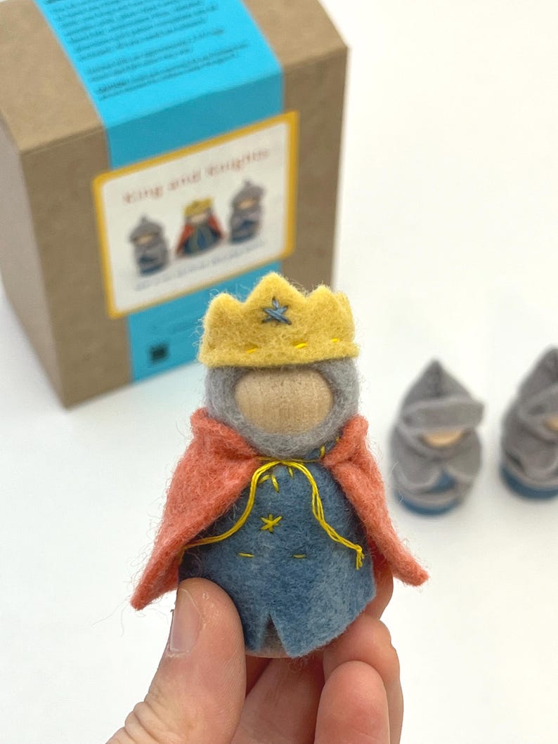 May include: A felt king figurine featuring a yellow crown, a grey hood, and a blue tunic with yellow stitching, draped in a coral-colored cape. The figure has a smooth wooden face and is part of a set including two grey knight figurines.