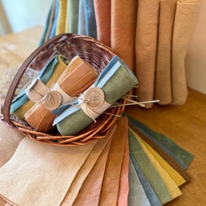 Op de afbeelding: Een verzameling kleurrijke vellen en rollen vilt in verschillende pasteltinten, waaronder geel, blauw, perzik en groen. Sommige viltrollen zijn vastgebonden met touw en labels. Het vilt wordt gepresenteerd in een rieten mand en op een houten oppervlak.