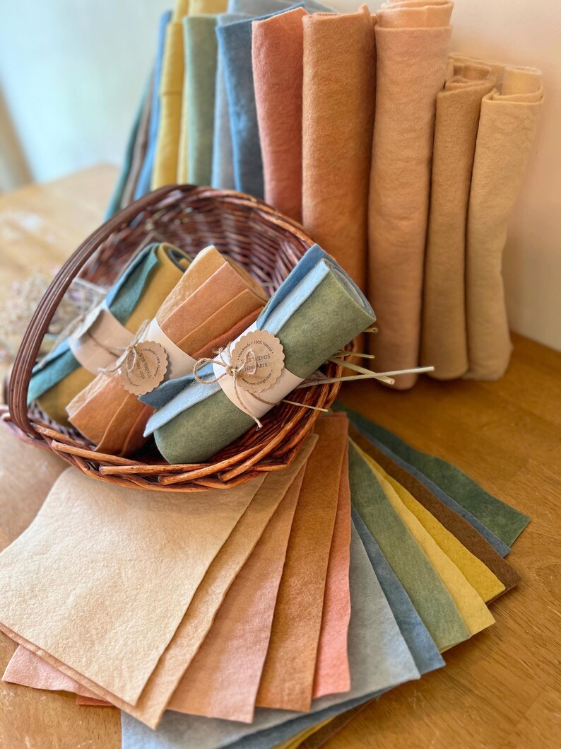 Op de afbeelding: Een verzameling kleurrijke vellen en rollen vilt in verschillende pasteltinten, waaronder geel, blauw, perzik en groen. Sommige viltrollen zijn vastgebonden met touw en labels. Het vilt wordt gepresenteerd in een rieten mand en op een houten oppervlak.