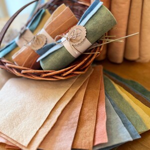 Op de afbeelding: Een verzameling kleurrijke vellen en rollen vilt in verschillende aardetinten. Rollen zijn gebundeld met touw en een label met de tekst "Sweet Things Arts". Het vilt is gerangschikt in een rieten mand en op een houten oppervlak.