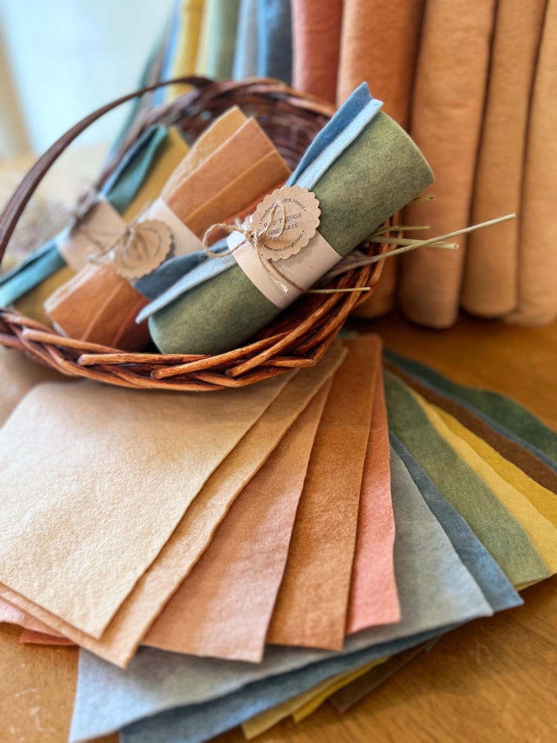 Op de afbeelding: Een verzameling kleurrijke vellen en rollen vilt in verschillende aardetinten. Rollen zijn gebundeld met touw en een label met de tekst "Sweet Things Arts". Het vilt is gerangschikt in een rieten mand en op een houten oppervlak.