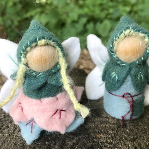 May include: Two small felted figures with round wooden heads, white wings, and dark green pointed hoods. One figure wears a light pink petal-shaped skirt with red stitching, while the other has a light blue body with a thin red waist tie.