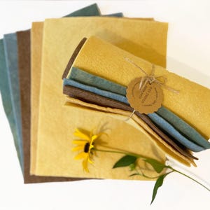 Op de afbeelding: Een verzameling vellen vilt in verschillende kleuren, waaronder tinten geel, blauw en bruin. Een bundel vilt is vastgebonden met touw en een label. Een gele bloem met groene bladeren is bij de vellen vilt geplaatst.