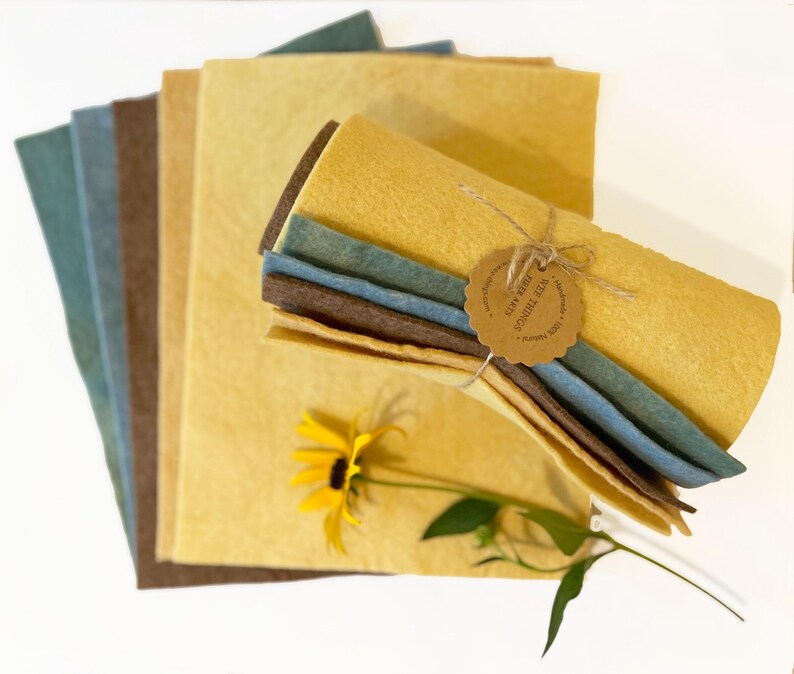 Op de afbeelding: Een verzameling vellen vilt in verschillende kleuren, waaronder tinten geel, blauw en bruin. Een bundel vilt is vastgebonden met touw en een label. Een gele bloem met groene bladeren is bij de vellen vilt geplaatst.