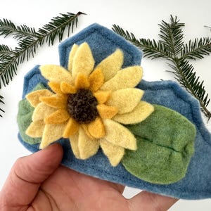 Op de afbeelding: Een handgemaakte blauwe vilten haarband met een grote vilten zonnebloem met gele bloemblaadjes en een donkerbruin centrum. Groene vilten bladeren zijn onder de bloem geplaatst. De haarband is versierd met een gestikt ontwerp.