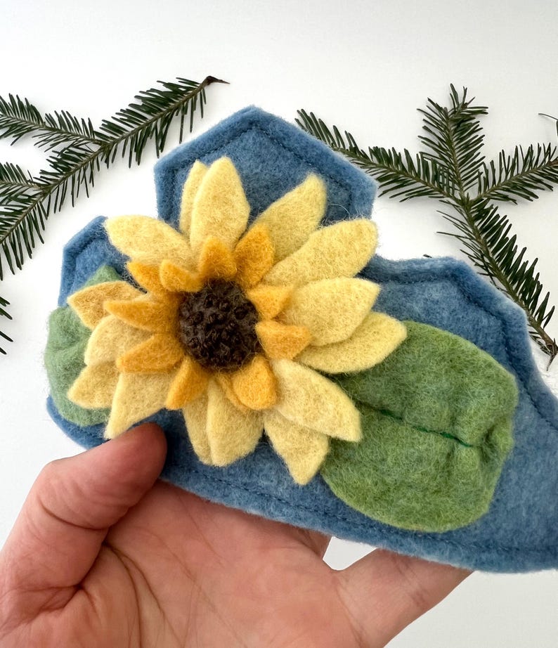 Op de afbeelding: Een handgemaakte blauwe vilten haarband met een grote vilten zonnebloem met gele bloemblaadjes en een donkerbruin centrum. Groene vilten bladeren zijn onder de bloem geplaatst. De haarband is versierd met een gestikt ontwerp.