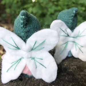 May include: Two small felted figures with pointed dark green caps and white fabric wings. The wings feature decorative green stitching details. A small portion of a light pink fabric base is visible beneath the white wings of the front figure.