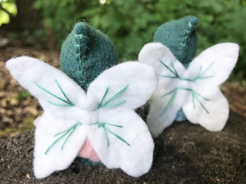 May include: Two small felted figures with pointed dark green caps and white fabric wings. The wings feature decorative green stitching details. A small portion of a light pink fabric base is visible beneath the white wings of the front figure.