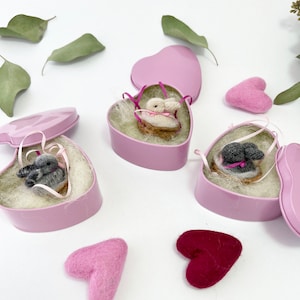 May include: Three heart-shaped, pink metal boxes, each containing a miniature knitted animal in a basket. The boxes are open, revealing the contents nestled in white fluff. Two felt hearts in shades of pink and red are scattered nearby. The background is white.