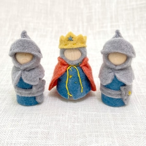 May include: Three small peg dolls with wooden heads. Two dolls wear grey felt hoods and blue felt bodies. The center doll wears a yellow felt crown, a red felt cape, and a blue felt body with yellow embroidered star details and yellow string ties.