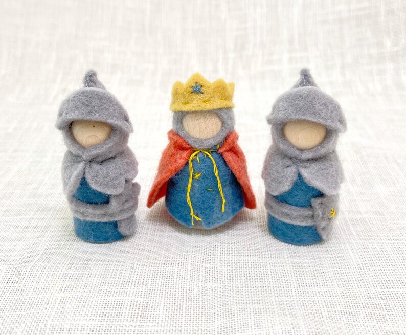 May include: Three small peg dolls with wooden heads. Two dolls wear grey felt hoods and blue felt bodies. The center doll wears a yellow felt crown, a red felt cape, and a blue felt body with yellow embroidered star details and yellow string ties.