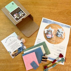 May include: A craft kit for making fairy folk dolls, including a brown cardboard box, a printed instruction sheet, two wooden doll heads, small rectangular pieces of felt in pink, blue, and green, a wooden spool with thread, and two embroidery floss skeins.