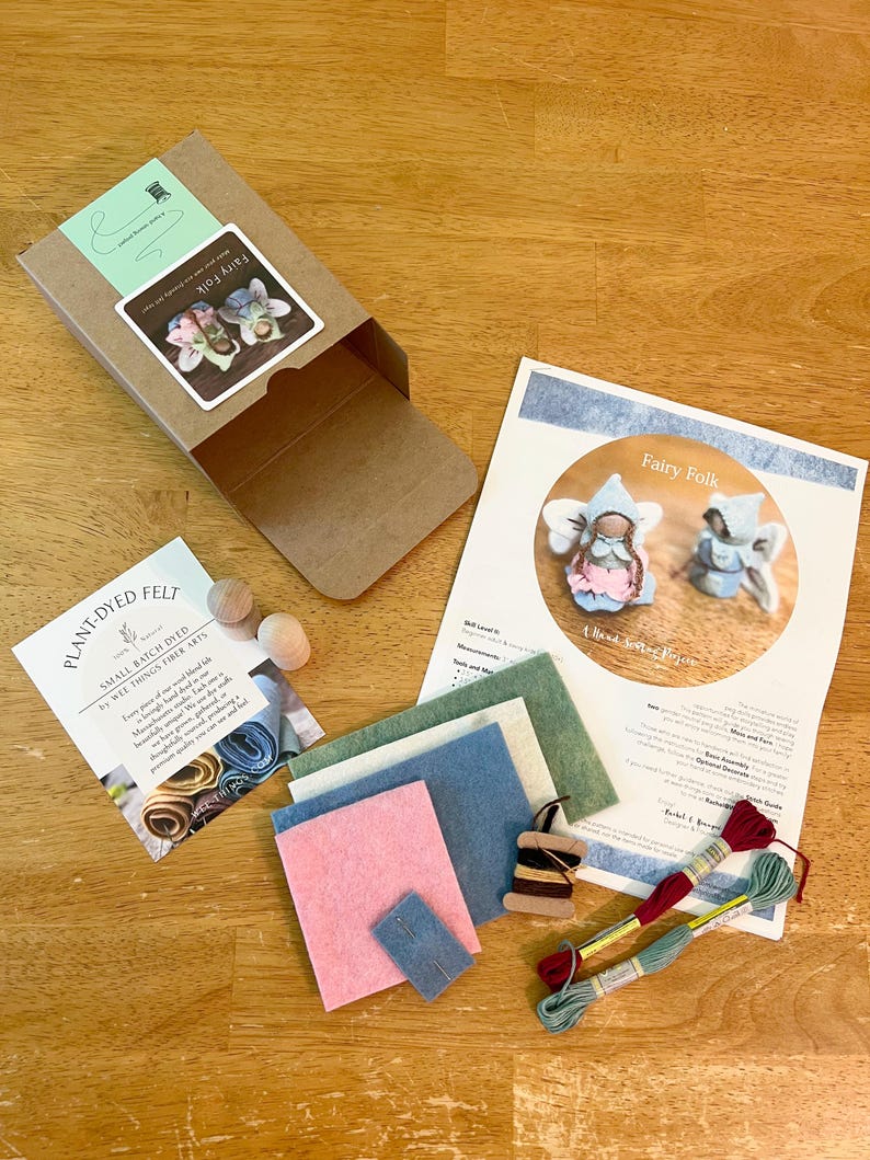 May include: A craft kit for making fairy folk dolls, including a brown cardboard box, a printed instruction sheet, two wooden doll heads, small rectangular pieces of felt in pink, blue, and green, a wooden spool with thread, and two embroidery floss skeins.