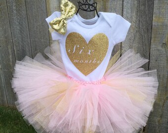May include: A white onesie with a gold glitter heart that says "Six Months" on it. The onesie is paired with a pink and gold tulle tutu.