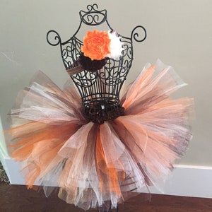 May include: A brown, orange, and white tulle tutu skirt with a brown ribbon waistband. The skirt is displayed on a black mannequin.