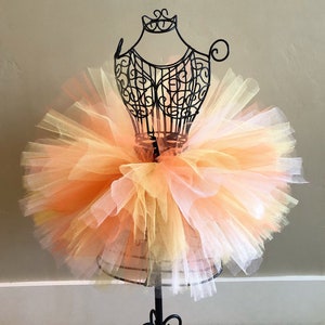 May include: A layered tutu skirt in shades of yellow, orange, and white is displayed on a black metal dress form. The tulle skirt is full and fluffy, with a gradient effect from light to darker colors.
