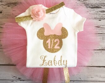 May include: Pink tulle tutu skirt with a gold glitter headband and a white onesie with a gold glitter Minnie Mouse design, the number 1/2 and the name "Eabby".