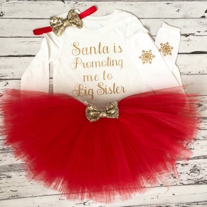 May include: A white long-sleeved shirt with the text "Santa is Promoting me to Big Sister" in gold glitter. The shirt has a gold glitter bow and snowflake embellishments. A red tulle tutu with a gold glitter bow is included.