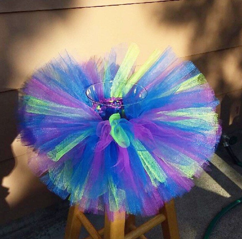 Puede incluir: Un tut&uacute; de tul colorido con capas verdes, azules y moradas. El tut&uacute; se est&aacute; utilizando como centro de mesa para un jarr&oacute;n.