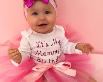 May include: A baby girl wearing a white onesie with the text "It's My Mommy Birthday" and a pink tulle tutu with a pink bow. She is wearing a pink headband with a large pink sequin bow.