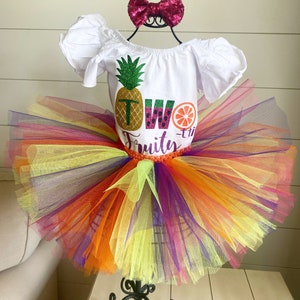 May include: A white tutu with rainbow tulle layers, a white shirt with a glittery pineapple, watermelon, and orange design, and the text "Two-ti-Fruity", and a pink bow.