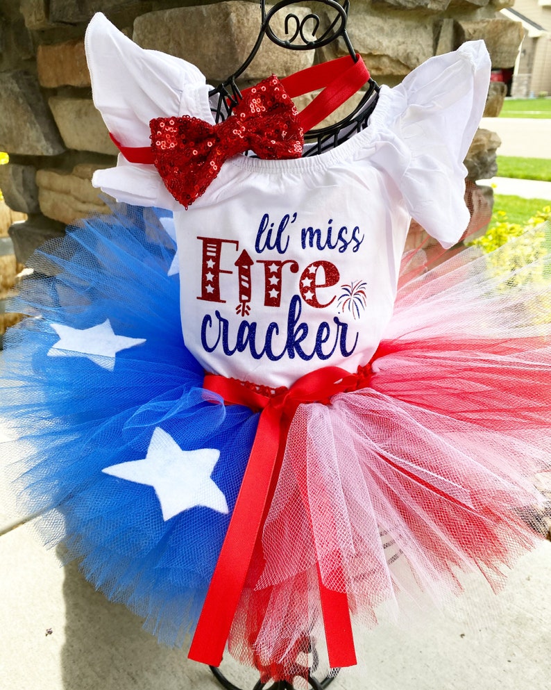K&ouml;nnte beinhalten: Ein wei&szlig;er Baby-Strampler mit dem Text "lil' miss Firecracker" in roten und blauen Buchstaben. Der Strampler wird mit einer roten Pailletten-Schleife und einem roten, wei&szlig;en und blauen T&uuml;ll-Tutu kombiniert.