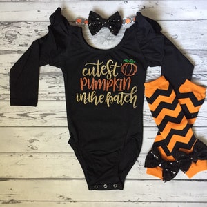 Może przedstawiać: Czarny, długorękawny body z napisem "cutest pumpkin in the patch" w złotym brokacie z pomarańczową grafiką dyni. Body ma falbankę na rękawach. Czarna kokardka z białymi kropkami jest na opasce. Pomarańczowe i czarne legginsy w chevron z czarną kokardką z cekinami są w zestawie.