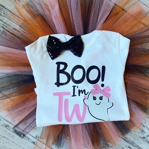 May include: A white onesie with the text "BOO! I'm TWO" in black and pink lettering. A white ghost with pink bow and a black bow tie are on the onesie. The onesie is on top of a brown and pink tulle tutu.
