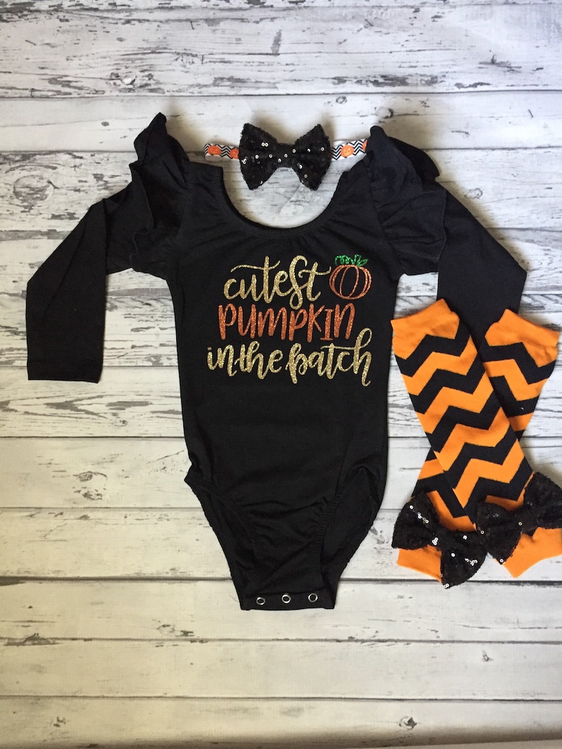 Może przedstawiać: Czarny, długorękawny body z błyszczącym złotym wzorem, na kt&oacute;rym widnieje napis "cutest pumpkin in the patch" i mały pomarańczowy obrazek dyni. Body ma falbanki na rękawach. Czarna kokardka z srebrnymi cekinami znajduje się na białej opasce. Pomarańczowo-czarne legginsy z chevronem i czarną kokardką z cekinami są dołączone.
