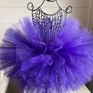 以下が含まれることがあります： 黒い金属製のマネキンに飾られた紫色のチュールチュチュ。チュチュはボリュームがありふわふわしていて、チュールが何層にも重なっています。